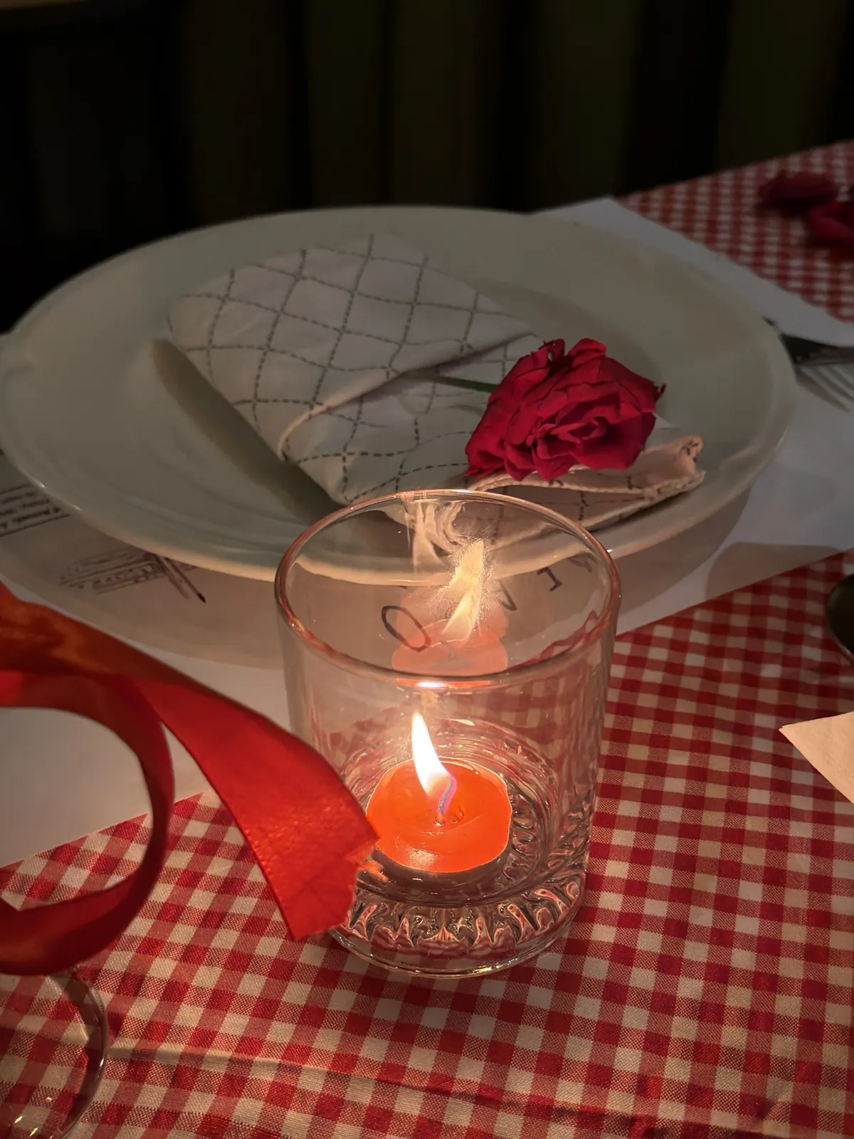 A lit candle on a red check tablecloth beside a folded napkin and a rose